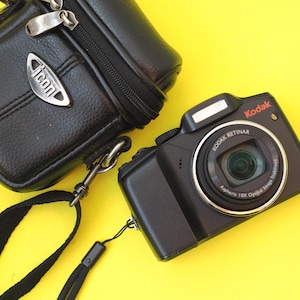 May include: A black Kodak digital camera with a Retinar lens and a black carrying case featuring an Icon logo. The camera has a lens ring labeled Kodak Retinar Aspheric 10X Optical Image Stabilized and a black wrist strap attached to the side.