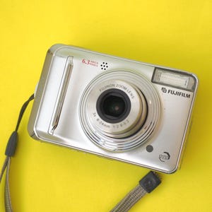 May include: A silver Fujifilm digital camera featuring a Fujinon zoom lens with 3x 8-24mm 1:2.8-5.3 specifications, a 6.3 megapixel sensor, a built-in flash, and a grey wrist strap attached to the side of the rectangular camera body.
