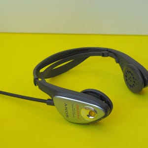 K&ouml;nnte beinhalten: Ein dunkelgraues Sony FM/AM-Radio-Headset mit umlaufendem Kopfb&uuml;gel. Das Ger&auml;t verf&uuml;gt &uuml;ber ein silberfarbenes Bedienfeld an der Seite mit einer kleinen orangefarbenen Sendeanzeige und einer d&uuml;nnen, flexiblen schwarzen Antenne auf der linken Seite.