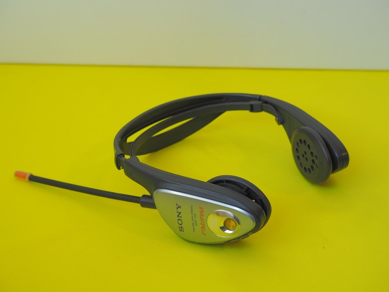 K&ouml;nnte beinhalten: Ein dunkelgraues Sony FM/AM-Radio-Headset mit umlaufendem Kopfb&uuml;gel. Das Ger&auml;t verf&uuml;gt &uuml;ber ein silberfarbenes Bedienfeld an der Seite mit einer kleinen orangefarbenen Sendeanzeige und einer d&uuml;nnen, flexiblen schwarzen Antenne auf der linken Seite.