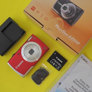May include: A red Canon PowerShot A2500 digital camera with a black battery charger, a black rechargeable battery pack, a black wrist strap, a product box featuring the camera image, and a Canon instructional booklet on a yellow background.