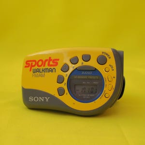 Puede incluir: Radio Sony Sports Walkman FM/AM amarilla y gris. La radio tiene una pantalla digital y varios botones para encendido, selecci&oacute;n de banda y emisoras preestablecidas. El texto "Sports Walkman FM/AM" est&aacute; impreso en la parte delantera.