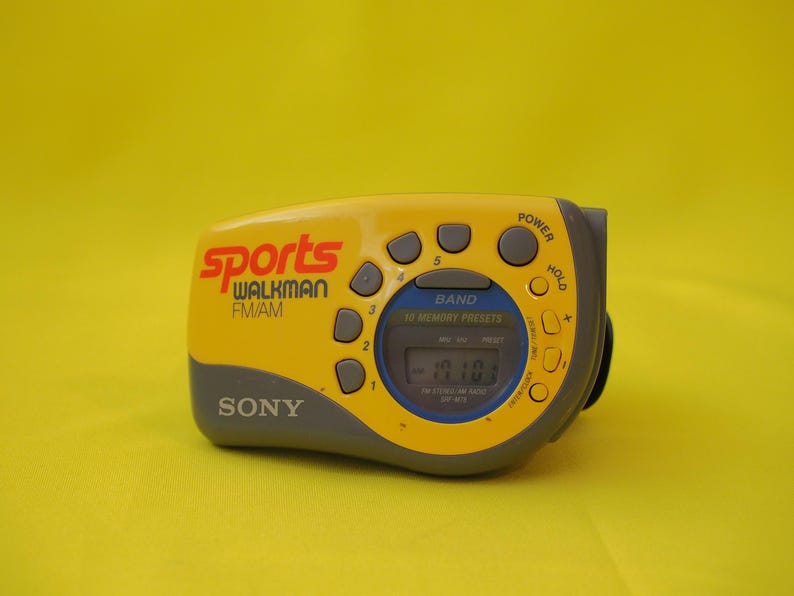 Puede incluir: Radio Sony Sports Walkman FM/AM amarilla y gris. La radio tiene una pantalla digital y varios botones para encendido, selecci&oacute;n de banda y emisoras preestablecidas. El texto "Sports Walkman FM/AM" est&aacute; impreso en la parte delantera.