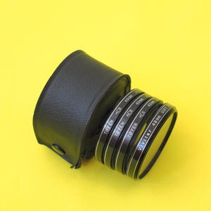 May include: A black leather-like circular carrying case next to a stack of four camera lens filters. The filters are labeled Tiffen HCE 49mm and Vivitar 49mm Soft. Each filter has a thin black metal frame with white printed text on the side.