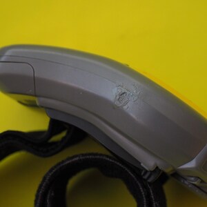 Puede incluir: Un dispositivo electr&oacute;nico gris y amarillo con una correa negra. El dispositivo tiene un &aacute;rea da&ntilde;ada en su costado. El fondo es de color amarillo liso.