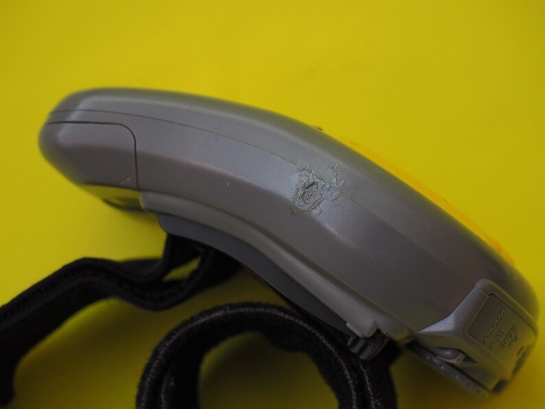 Puede incluir: Un dispositivo electr&oacute;nico gris y amarillo con una correa negra. El dispositivo tiene un &aacute;rea da&ntilde;ada en su costado. El fondo es de color amarillo liso.