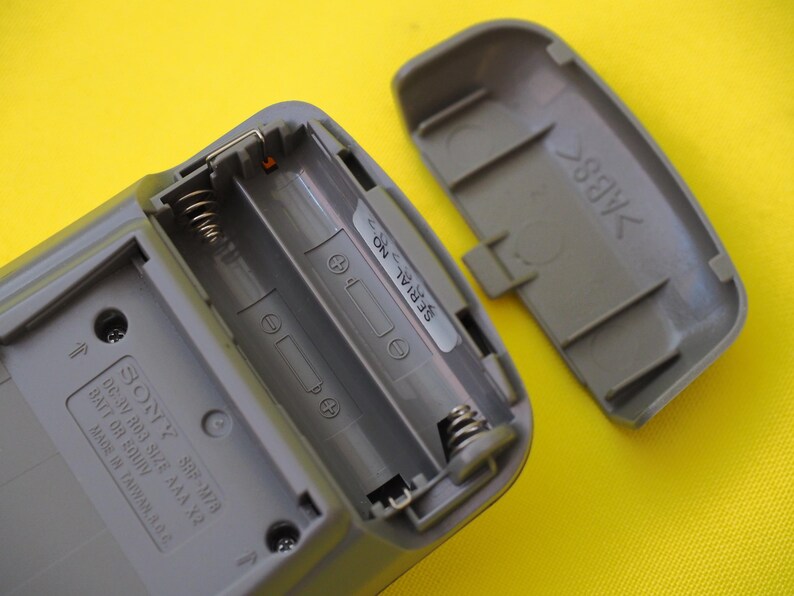 Puede incluir: Compartimento de pilas de pl&aacute;stico gris abierto de un dispositivo Sony, que revela dos ranuras para pilas AAA con muelles. La tapa separada muestra el interior. El texto "SERIAL NO." est&aacute; impreso en una pegatina.