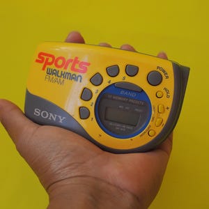 Puede incluir: Radio Sony Sports Walkman FM/AM amarilla y gris. El dispositivo tiene m&uacute;ltiples botones, una pantalla digital y el texto "Sports Walkman FM/AM" en rojo y blanco. La radio se sostiene en una mano sobre un fondo amarillo.