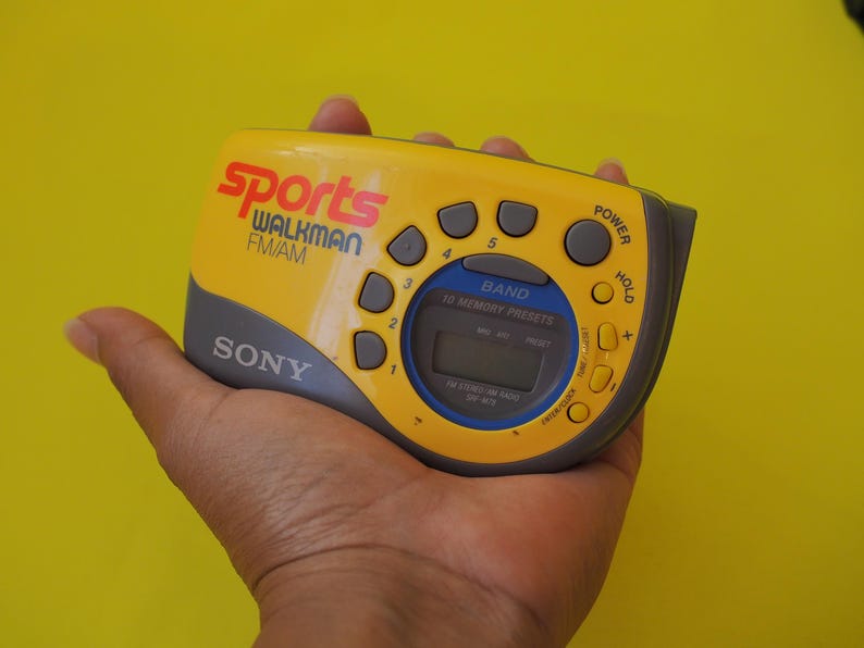 Puede incluir: Radio Sony Sports Walkman FM/AM amarilla y gris. El dispositivo tiene m&uacute;ltiples botones, una pantalla digital y el texto "Sports Walkman FM/AM" en rojo y blanco. La radio se sostiene en una mano sobre un fondo amarillo.