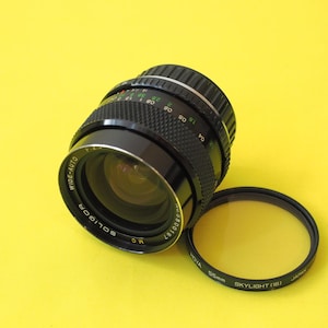 May include: A black Soligor Wide-Auto camera lens with a yellow filter. The lens has measurement markings and the text "SOLIGOR MC" and "WIDE-AUTO". The filter is labeled "55mm SKYLIGHT (1B)". The lens and filter are on a yellow background.