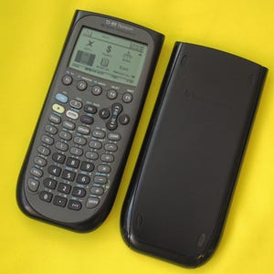 May include: A dark gray TI-89 Titanium graphing calculator with its protective cover. The calculator has numerous buttons and a display screen showing various icons. The cover is black and rests beside the calculator on a yellow surface.