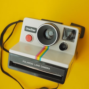 Peut inclure: Un appareil photo Polaroid OneStep Land Camera blanc avec un objectif noir et une bande arc-en-ciel. Le bouton est rouge et le texte "Polaroid Land Camera" est imprimé. Une sangle noire est attachée.