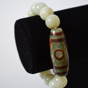 Puede incluir: Un brazalete hecho de cuentas de jade verde claro con una cuenta de ágata marrón y verde en el centro.