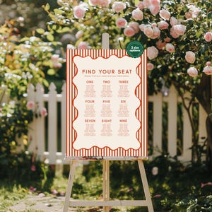 May include: A cream-colored seating chart with red striped borders and the text "FIND YOUR SEAT." The chart is divided into nine sections, each labeled with a number. A green circle with the text "2 size options" is in the upper right corner. The chart is displayed on a wooden easel.
