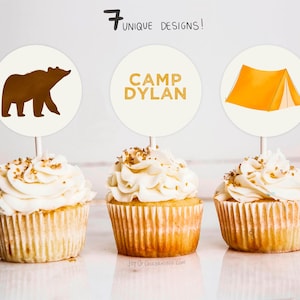 Op de afbeelding: Drie cupcakes met witte frosting en gouden sprinkles, bedekt met ronde papieren cupcake toppers. De toppers tonen een bruine beer, de tekst "CAMP DYLAN" en een gele tent.