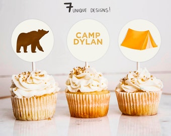 May include: Three cupcakes with white frosting and gold sprinkles topped with round paper cupcake toppers. The toppers feature a brown bear, the text "CAMP DYLAN", and a yellow tent.