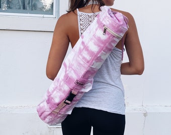 May include: A pink and white tie-dye yoga mat carrier with a zippered pocket and adjustable strap. The carrier has the word "The Journey" printed on the strap.