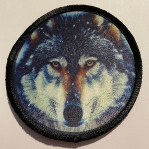Puede incluir: Un parche redondo con borde negro que muestra una imagen de primer plano de la cara de un lobo. El lobo tiene un pelaje gris azulado, ojos marrones y una nariz negra.