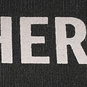 May include: A black rectangular patch with the words "SHE HER HERS" printed in white, bold, capital letters. The text is centered and the background has a textured appearance.