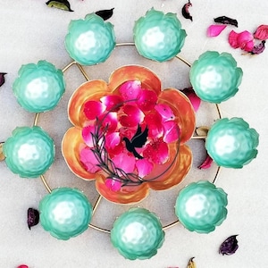 以下が含まれることがあります： 丸い天板と8つのターコイズブルーのボウルが円形に配置された、金色の金属製の装飾的なスタンド。中央のボウルは、ピンクのバラの花びらで満たされた、より大きなオレンジ色のボウルです。