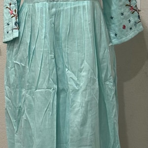 May include: A light blue long-sleeved dress featuring a notched neckline and a gathered waist. The bodice and sleeves are decorated with embroidered floral patterns in pink, red, and dark blue, including a central bird motif on the chest.