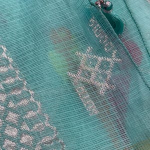 May include: Close-up of a sheer, turquoise fabric with a silver geometric pattern. A small turquoise tassel with a decorative silver bead is attached to the fabric. The fabric has a delicate, woven texture and a subtle sheen.