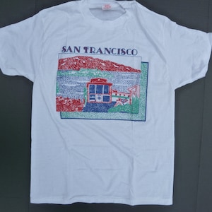 May include: White t-shirt featuring a graphic print of a red, blue and green illustration of a cable car in San Francisco. The text "SAN FRANCISCO" is printed above the illustration.
