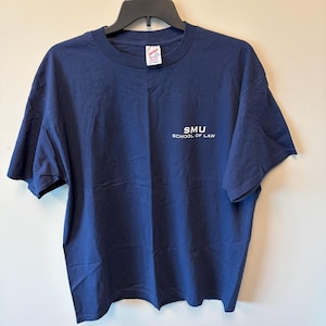 May include: A navy blue short-sleeved t-shirt featuring the white text SMU SCHOOL OF LAW printed on the right chest area. The shirt has a crew neckline and is displayed on a black plastic hanger against a plain, light-coloured background.