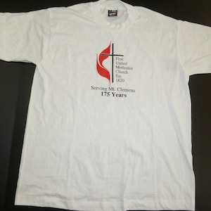 May include: White t-shirt with a red and black logo that says "First United Methodist Church Est. 1820 Serving Mt. Clemens 175 Years"