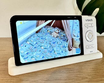 Op de afbeelding: Een VTech babyfoon met een witte behuizing en een zwart scherm dat een live videobeeld toont van een kinderbed met blauw beddengoed met patronen, een knuffeldier en een hemeltje. Het apparaat heeft een navigatieknop en diverse bedieningstoetsen.
