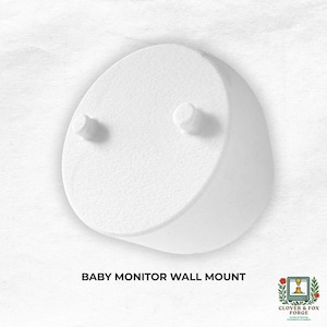 May include: A white baby monitor wall mount with two small cylindrical pegs. The mount has a curved, rounded shape. The text "BABY MONITOR WALL MOUNT" is below the mount. The Clover & Fox Forge logo is in the bottom right corner.
