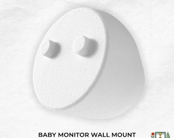 May include: A white, rounded, egg-shaped baby monitor wall mount with two small, circular protruding pegs on the front surface. The text "BABY MONITOR WALL MOUNT" is printed in black below the object, alongside a small logo for Clover & Fox Forge.