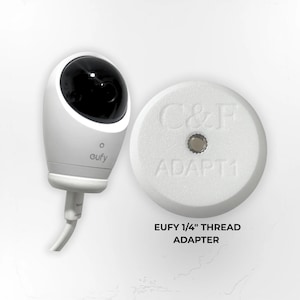 Może przedstawiać: Biała kamera bezpieczeństwa Eufy i biały adapter. Kamera ma czarny obiektyw i nadruk "eufy". Adapter jest okrągły z napisem "C&F ADAPT1" i metalową śrubą. Poniżej znajduje się tekst "EUFY 1/4" THREAD ADAPTER".