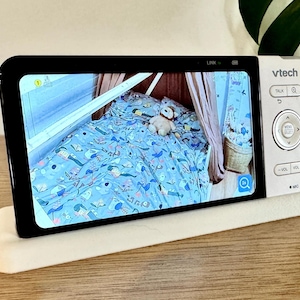 May include: A VTech baby monitor with a white frame and black screen displaying a nursery scene with a blue patterned bedspread and a stuffed animal. The device features a circular navigation pad, volume buttons, and a talk button on the right side.