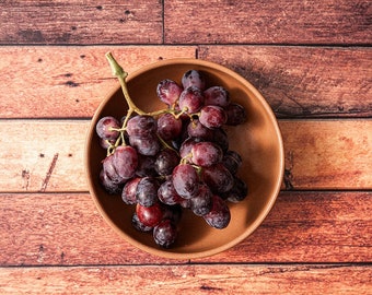 May include: A bowl of red grapes on a wooden surface. The grapes are arranged in a cluster and have a dark purple hue.