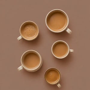 Könnte beinhalten: Draufsicht auf fünf beige Keramikbecher, gefüllt mit Kaffee, auf braunem Hintergrund. Die Becher sind unterschiedlich groß und haben ein schlichtes Design. Der Kaffee ist hellbraun, und die Anordnung ist ansprechend.