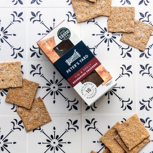 May include: A box of Peter's Yard Fig & Spelt Sourdough Crackers on a white tiled surface with black floral designs. The box is red and black with a white logo and text. The crackers are square and brown.