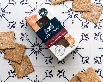 May include: A box of Peter's Yard Fig & Spelt Sourdough Crackers on a white tiled surface with black floral designs. The box is red and black with a white logo and text. The crackers are square and brown.
