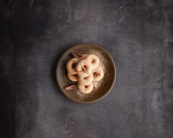 May include: A brown plate with a pile of small, round, sugared doughnuts. The doughnuts are arranged in a pile with a few cinnamon sticks on top.