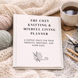 Può includere: Un'agenda con copertina rigida intitolata "The Cozy Knitting & Mindful Living Planner" con il testo "Uno spazio delicato per i tuoi progetti, le tue routine e le tue giornate lente". L'agenda è su una coperta di maglia color crema, con una tazza di caffè e accessori per lavorare a maglia.