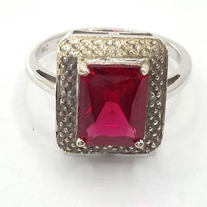 May include: A silver ring featuring a large, rectangular, deep red gemstone. The gemstone is set in a square frame with a textured border. The ring band is smooth and shiny.
