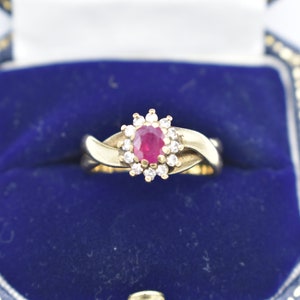 Könnte beinhalten: Ein goldener Ring mit einem ovalen Rubin in der Mitte, umgeben von kleinen Diamanten. Der Ring hat ein verdrehtes Goldband. Präsentiert in einer blauen Samt-Schmuckschatulle.