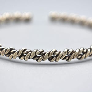 May include: A silver bangle bracelet with a braided design. The bracelet is made of a series of interlocking loops that create a rope-like effect.