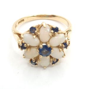 May include: A gold ring with a floral design. The flower is made up of oval white stones and small, round blue stones. The ring has a polished finish. The ring is set against a white background.