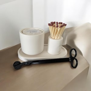 May include: A white ceramic candle holder with a lid, a small white ceramic cup filled with matches, and a black candle wick trimmer on a white tray. The matches have brown tips. The items are on a light brown surface.