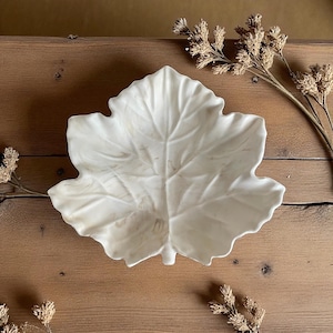 May include: A white, leaf-shaped decorative bowl with detailed veining. The bowl is made of a glossy material and sits on a wooden surface. Dried floral accents are arranged around the bowl, adding a natural touch.