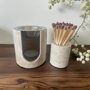 May include: A marble-effect candle holder with a silver bowl and a small opening for a tea light. Beside it is a matching cylindrical container filled with wooden matches with maroon tips. The items are on a dark wooden surface.