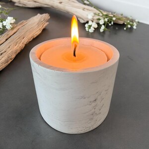 May include: A lit candle in a marbled grey concrete holder. The candle has a bright orange flame. Decorative driftwood and white flowers are in the background. The candle holder is approximately 7.6 cm in diameter.