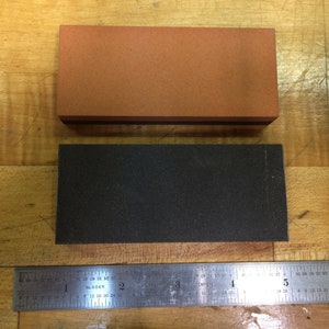 May include: Two rectangular sharpening stones, one brown and one black, are placed on a wooden surface. A metal ruler is positioned below the stones.