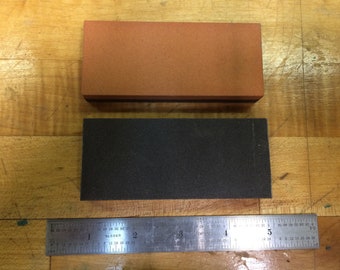 May include: Two rectangular sharpening stones, one brown and one black, are placed on a wooden surface. A metal ruler is positioned below the stones.